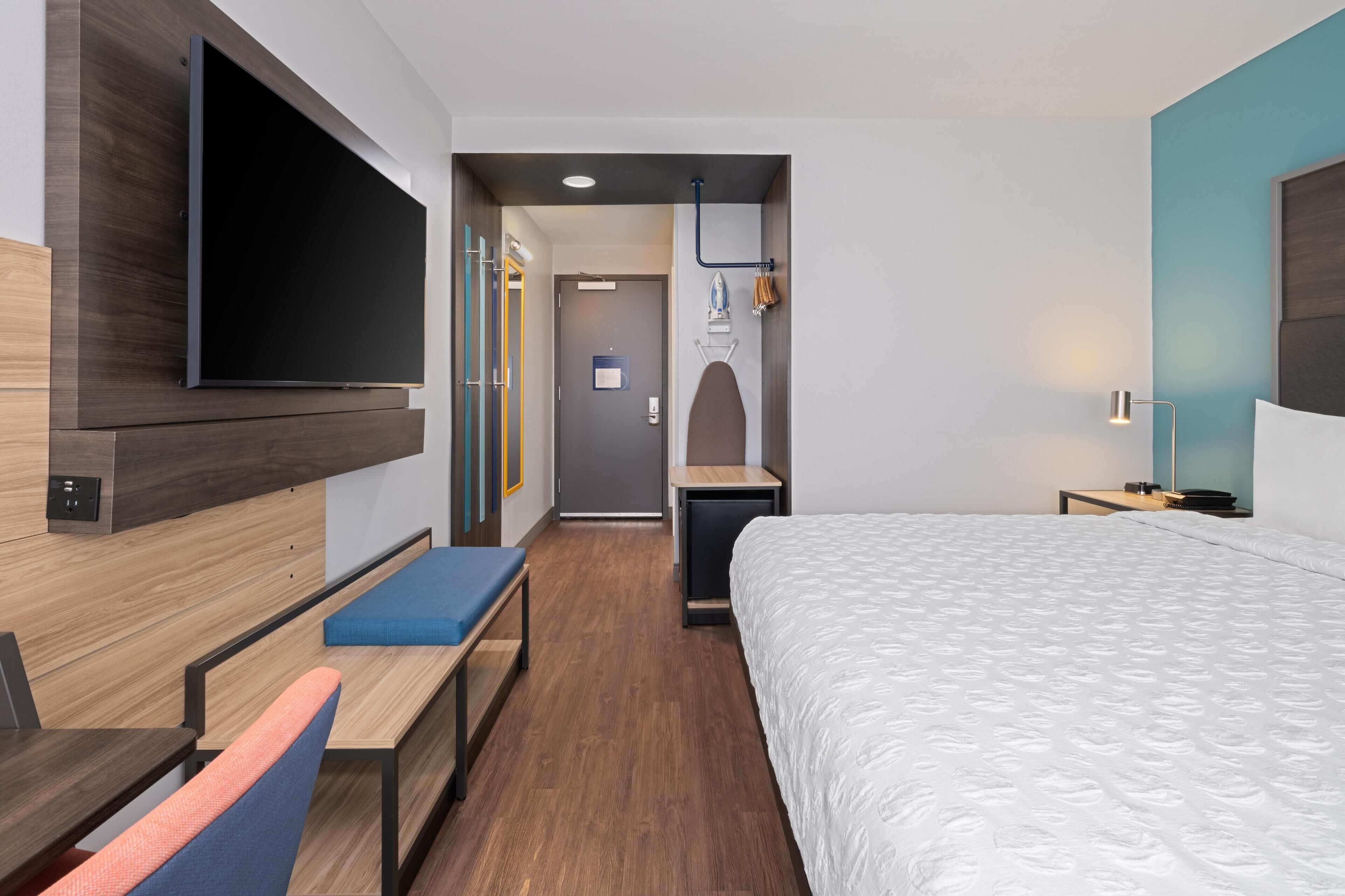 Quarto de hotel moderno com uma cama grande, uma TV de tela plana na parede, uma escrivaninha com cadeira e um banco.