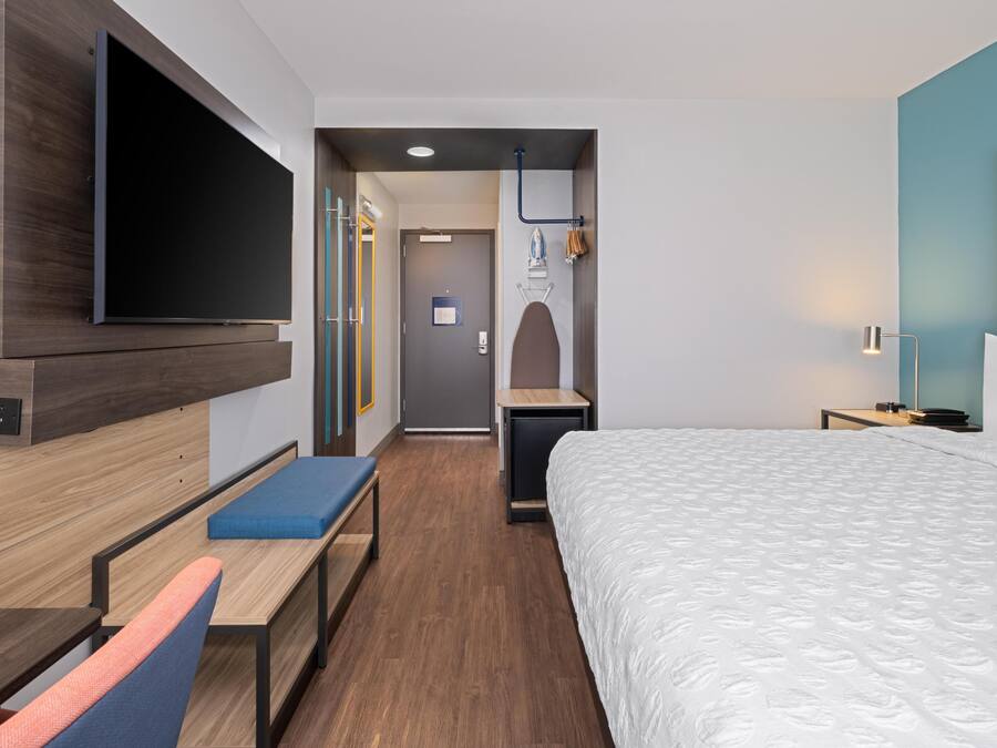 Quarto de hotel moderno com uma cama grande, uma TV de tela plana na parede, uma escrivaninha com cadeira e um banco.