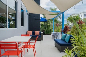 An outdoor patio area with a white table and orange chairs, a blue sofa with teal cushions, and a large canopy providing shade.