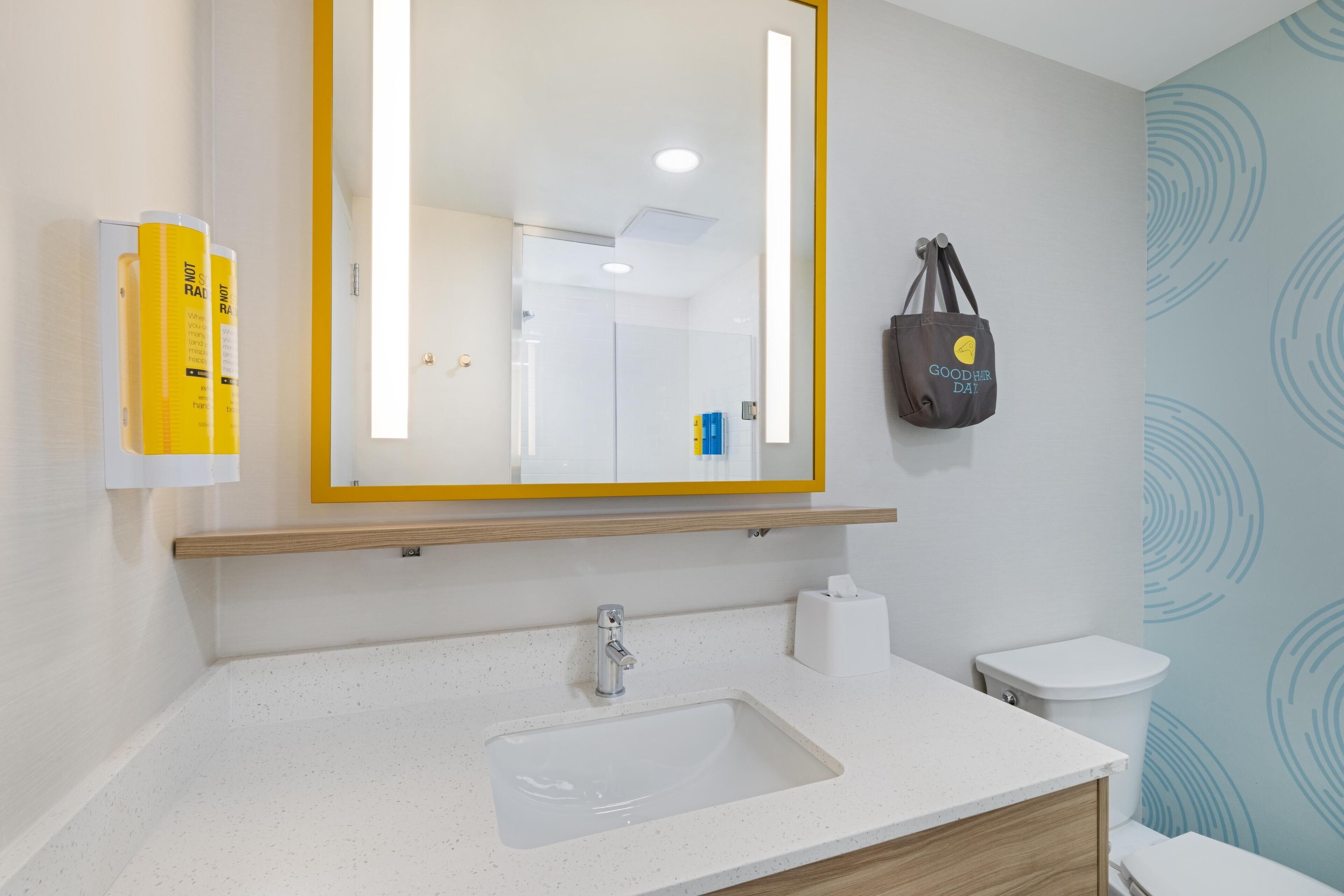 A modern bathroom with a white sink, a mirror with a yellow frame, a towel dispenser, and a toilet.