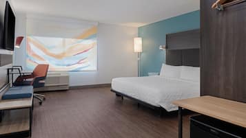 A hotel room with a bed, a desk, a chair, a TV, and a large abstract painting on the wall.