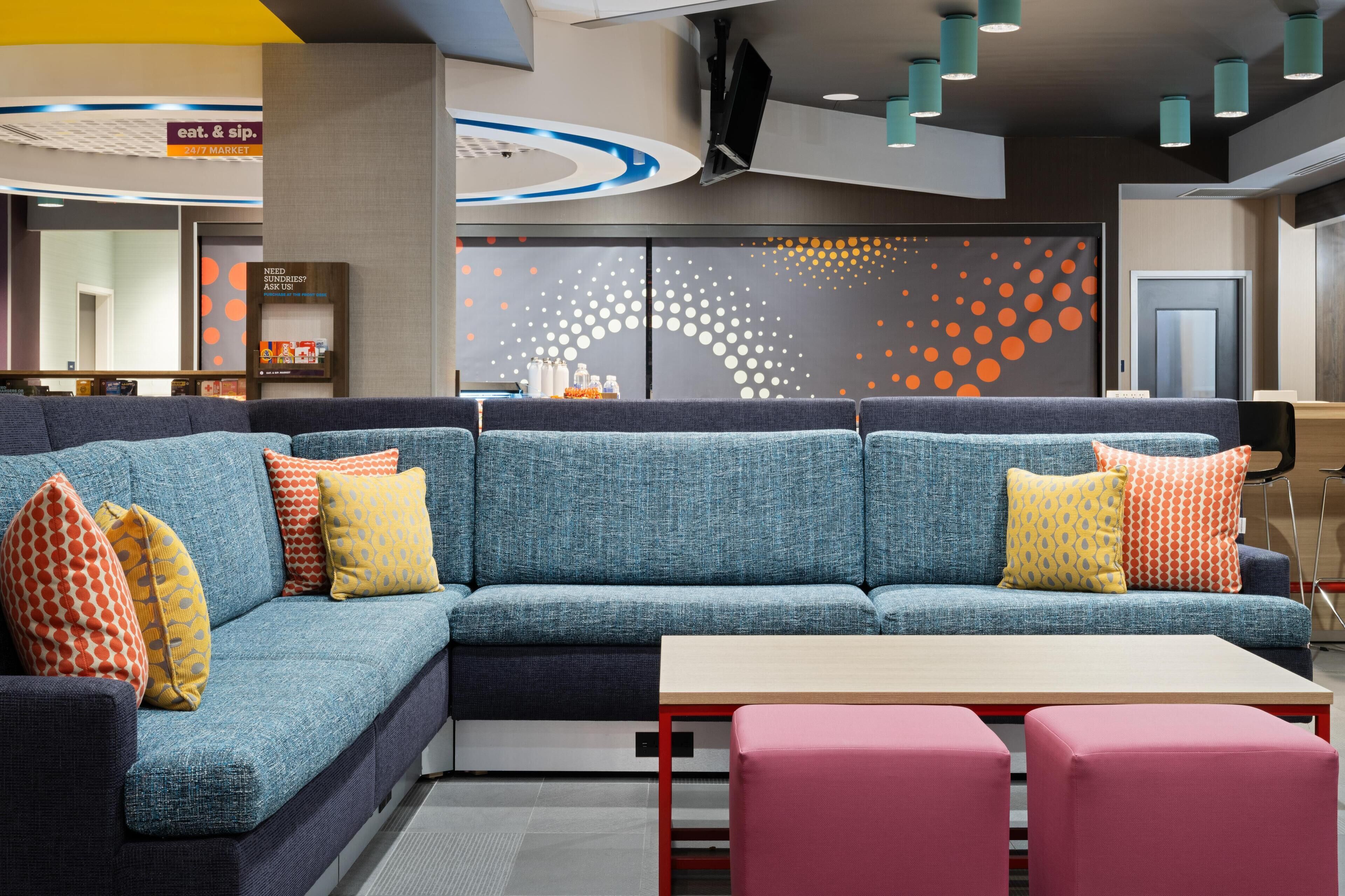 Zona relax moderna con un divano componibile blu, un tavolino e pouf rosa. Un cartello segnala un mercato aperto 24 ore su 24 e la necessità di richiedere assistenza.
