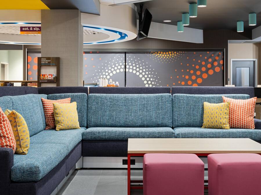 Een moderne zithoek met een blauwe modulaire bank, een klein tafeltje en roze poufs. Een bordje wijst op een 24/7 markt en vraagt om hulp.