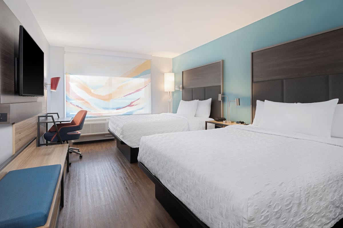 Une chambre d’hôtel avec deux lits, un bureau, une chaise, une télévision et un grand tableau abstrait accroché au mur.