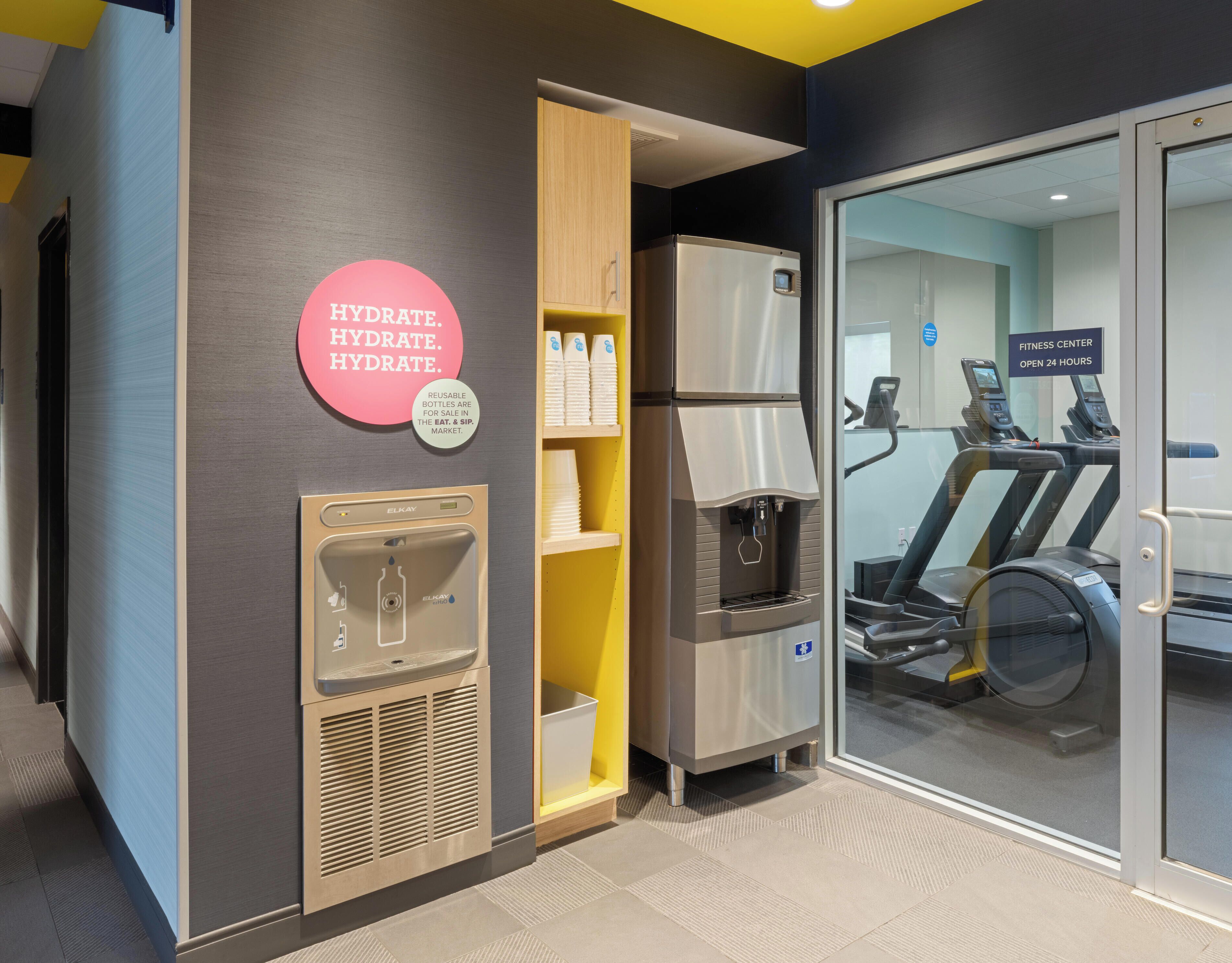 A hydration station with a water dispenser and reusable bottles, a fitness center with treadmills, and a sign indicating it's open 24 hours.