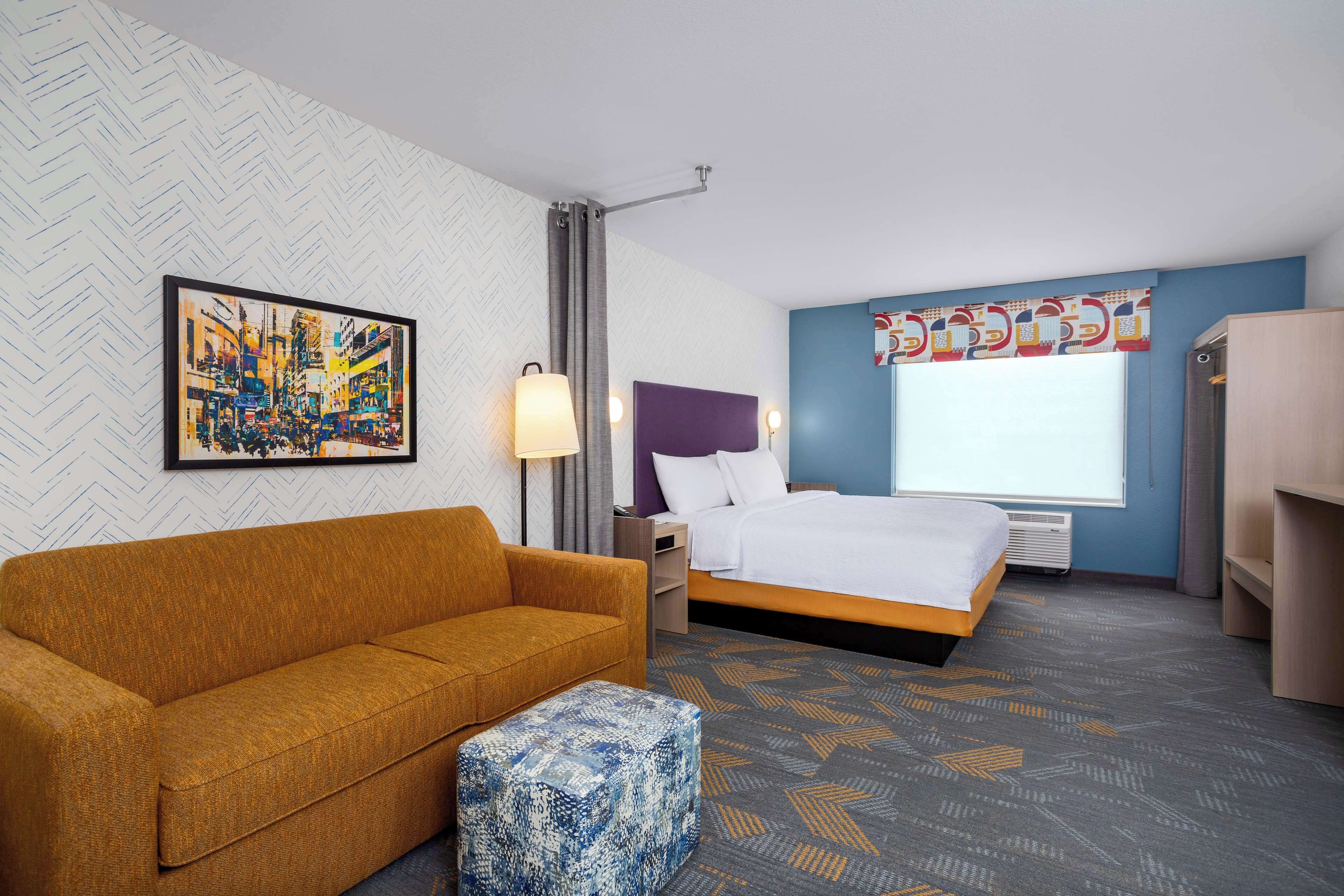 A hotel room with a sofa, a bed, a nightstand, a lamp, a window with curtains, and a wall with a herringbone pattern.