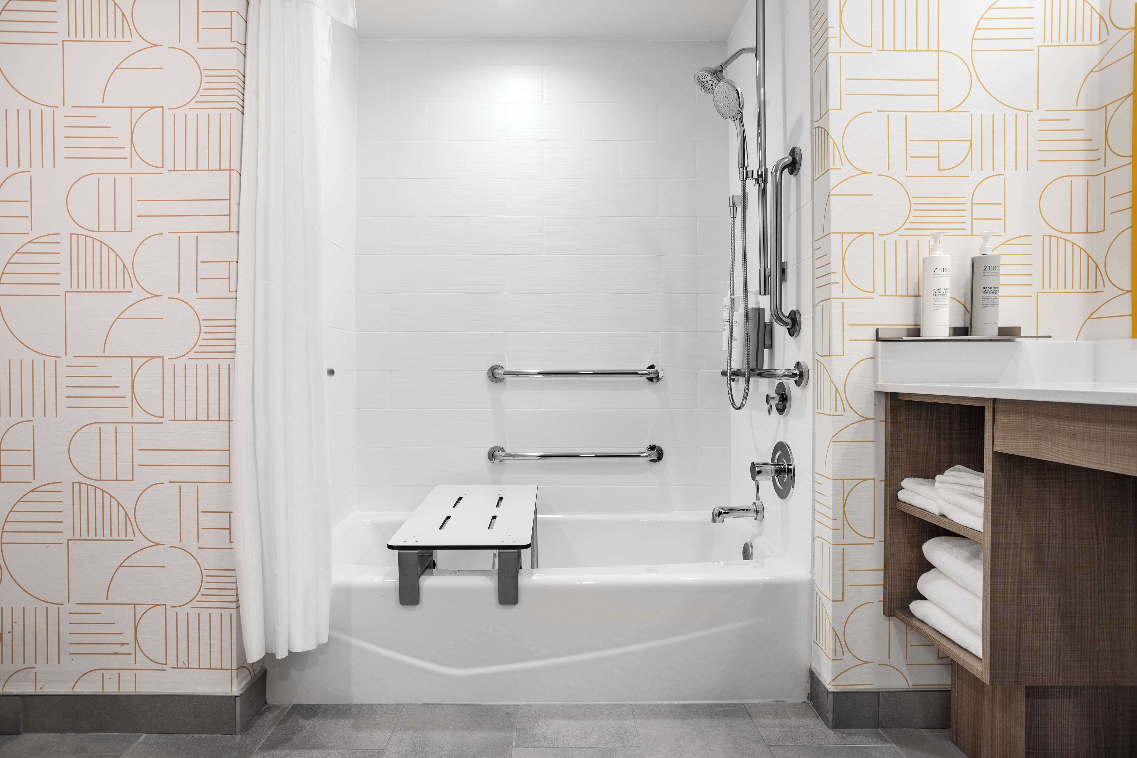 A modern bathroom with a white bathtub, a shower area, a wooden vanity with a towel rack, and a geometric patterned wall.