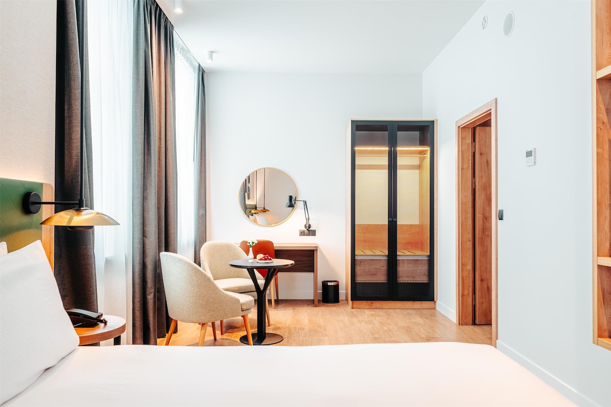 A hotel room with a bed, a desk, a chair, a lamp, a round mirror, and a view of a balcony.