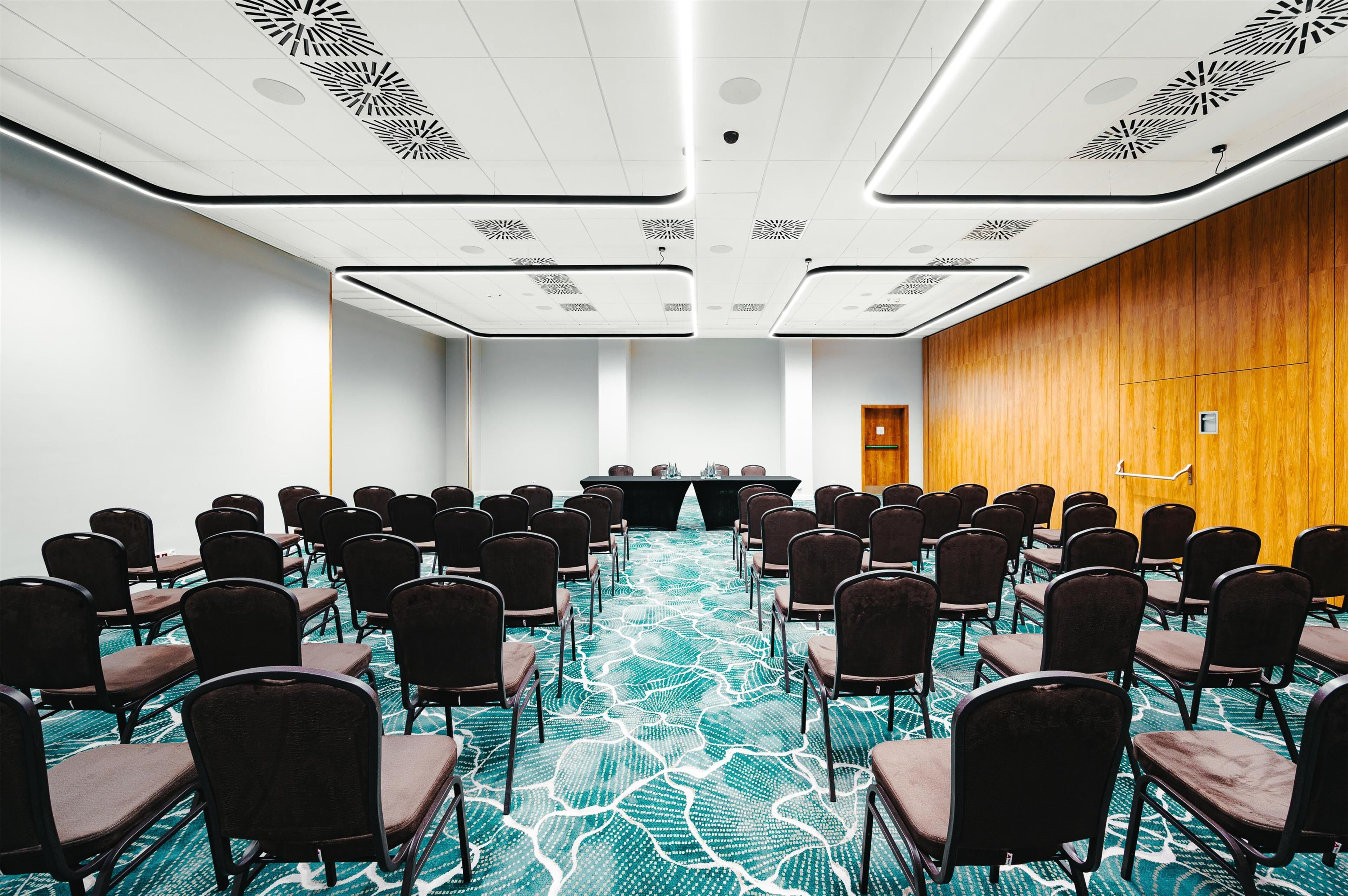Uma sala de conferências com fileiras de cadeiras voltadas para uma mesa comprida, carpete estampado e parede de painéis de madeira.