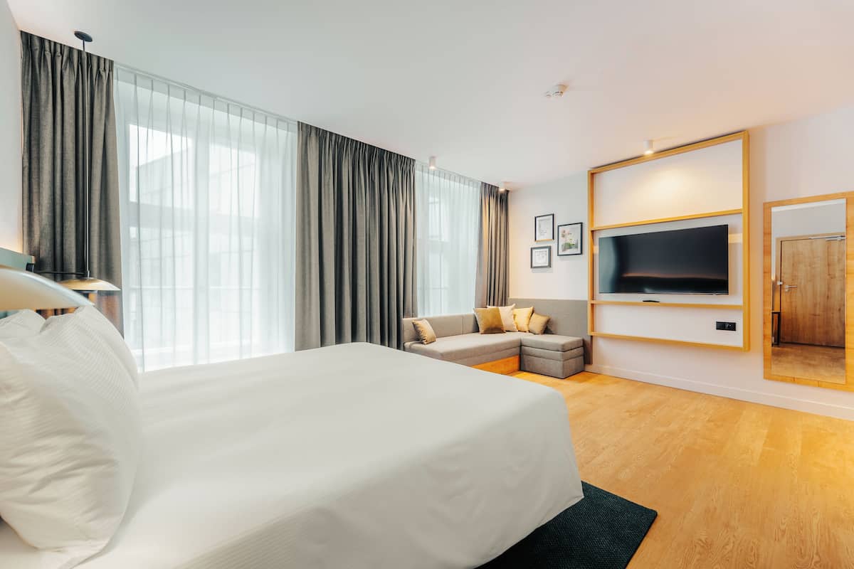 A modern hotel room with a large bed, a sofa, a TV mounted on the wall, and a large window with curtains.
