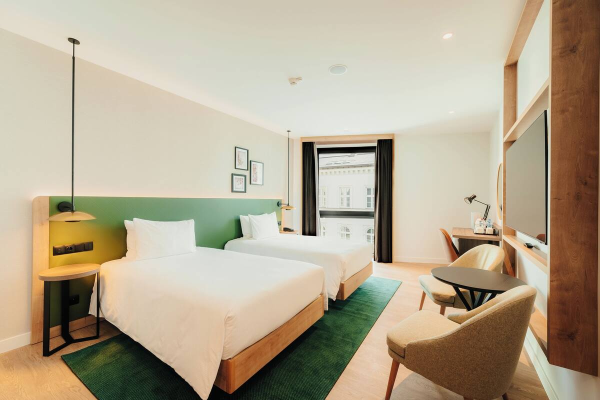 A hotel room with two beds, a desk, a chair, a TV, and a window with a view of buildings.