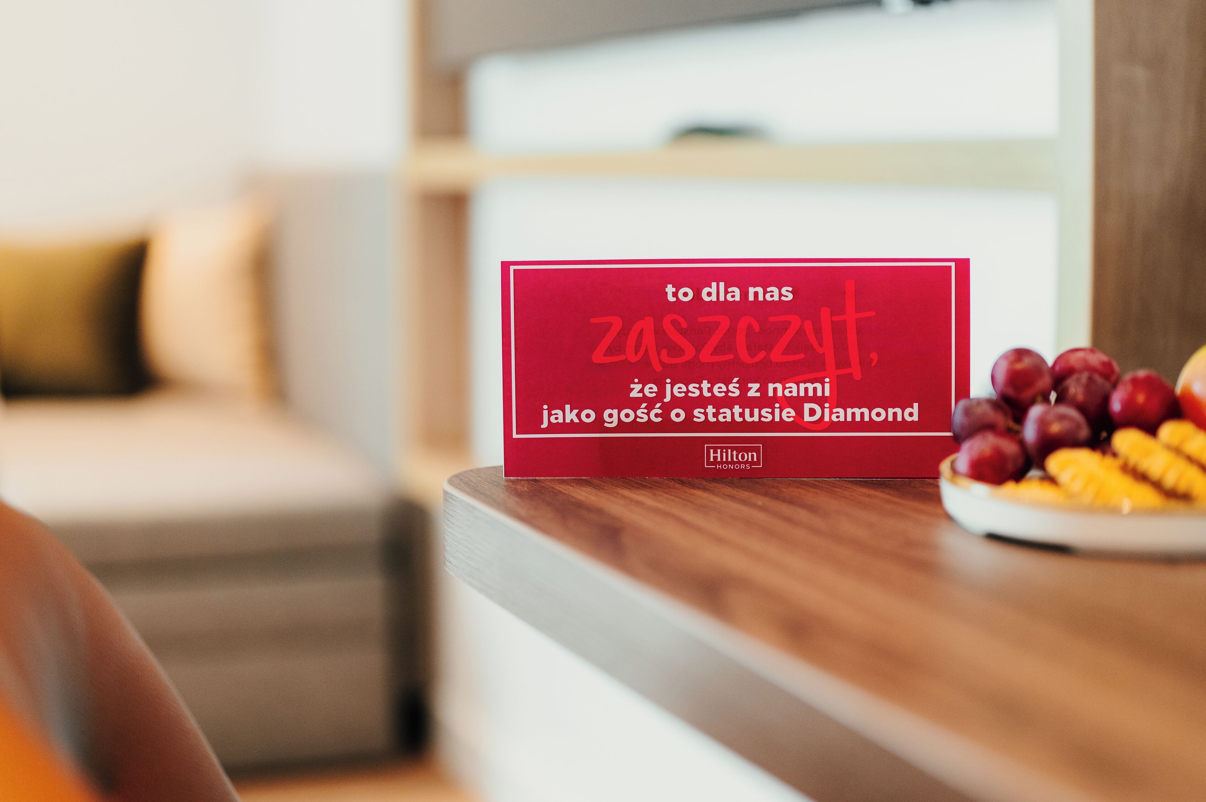 A red sign with text in a foreign language, placed on a wooden surface next to a plate of fruit.