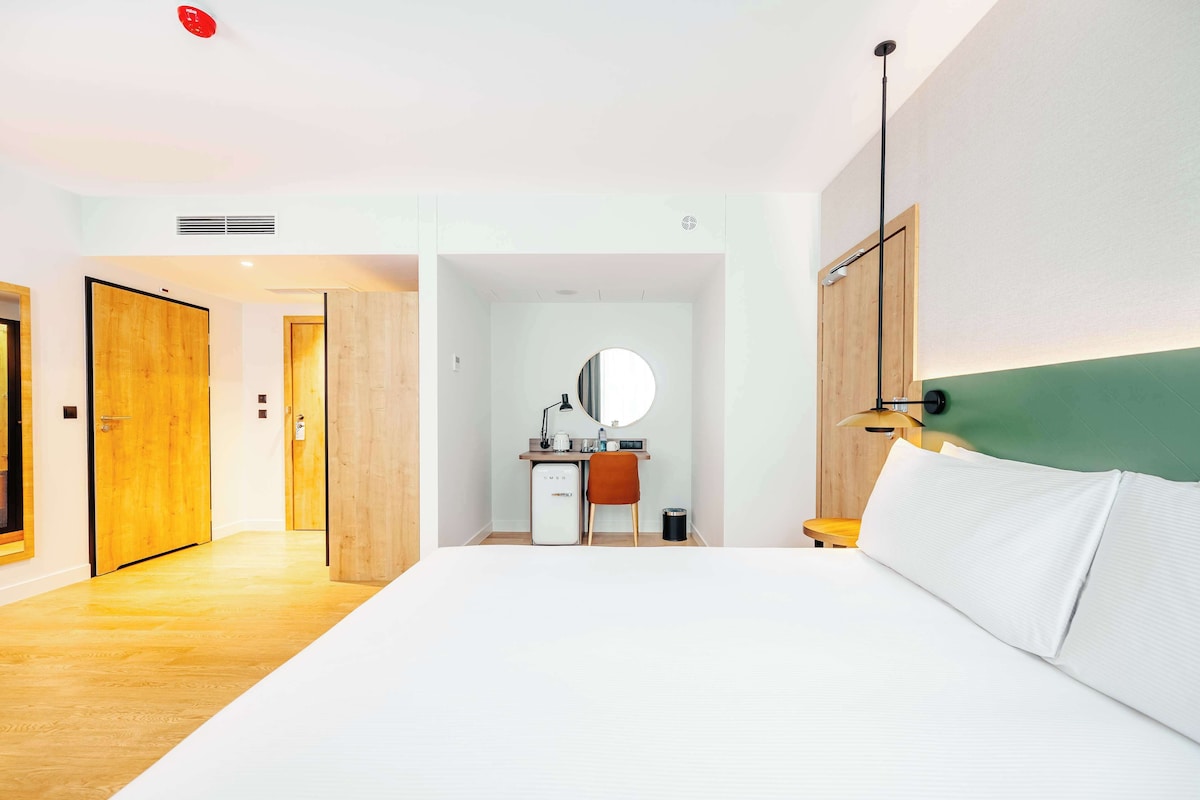 A hotel room with a large bed, a desk with a mirror, a small fridge, and a wooden floor.