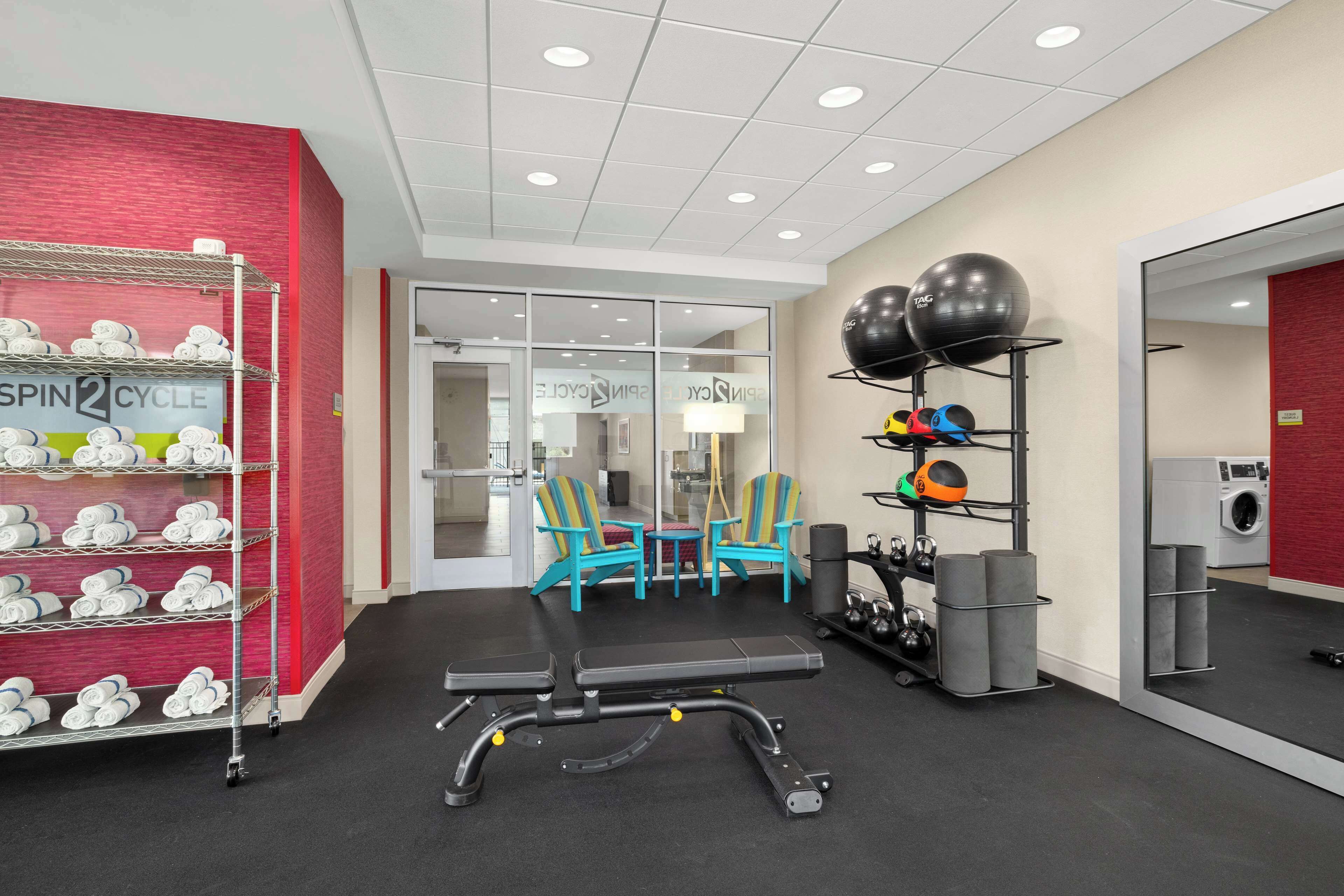 Une salle de sport équipée de matériel d’exercice, d’un banc, d’un porte-balles et d’une étagère murale où sont rangées des serviettes.