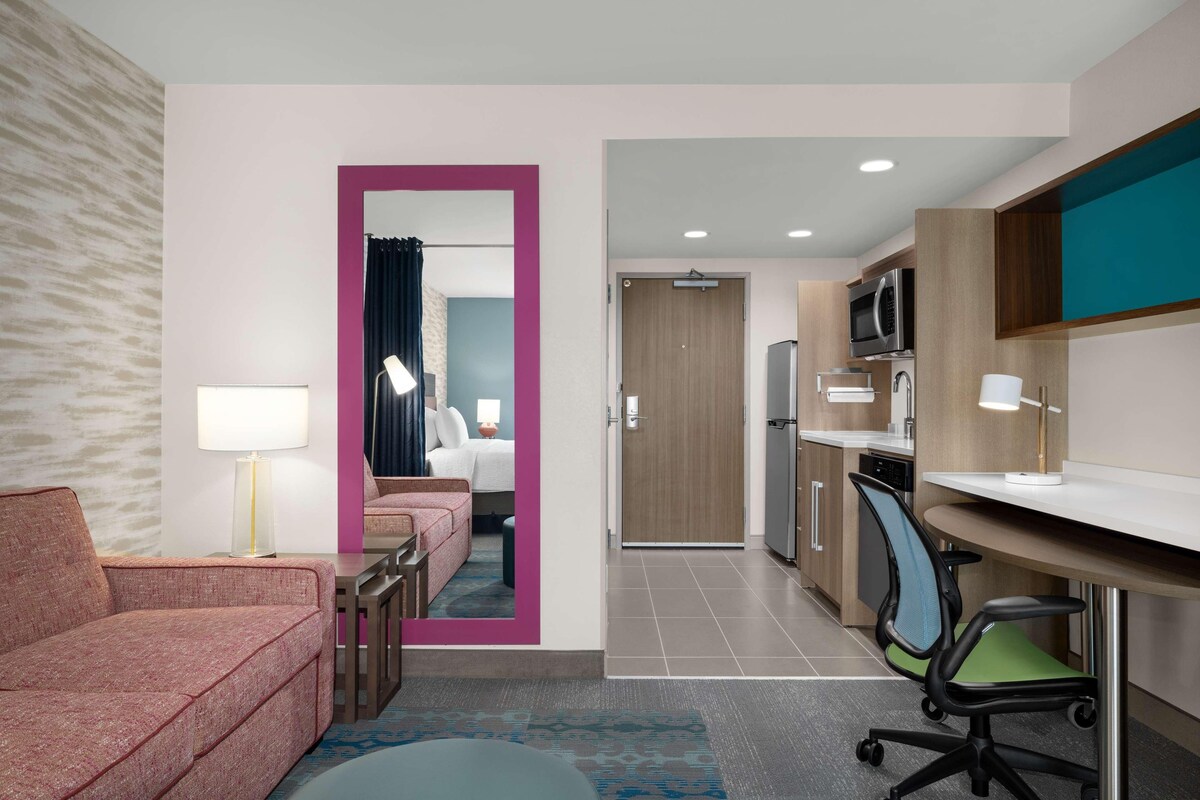 A modern hotel room with a pink-framed mirror, a sofa, a desk with a computer, a microwave, and a small kitchenette.