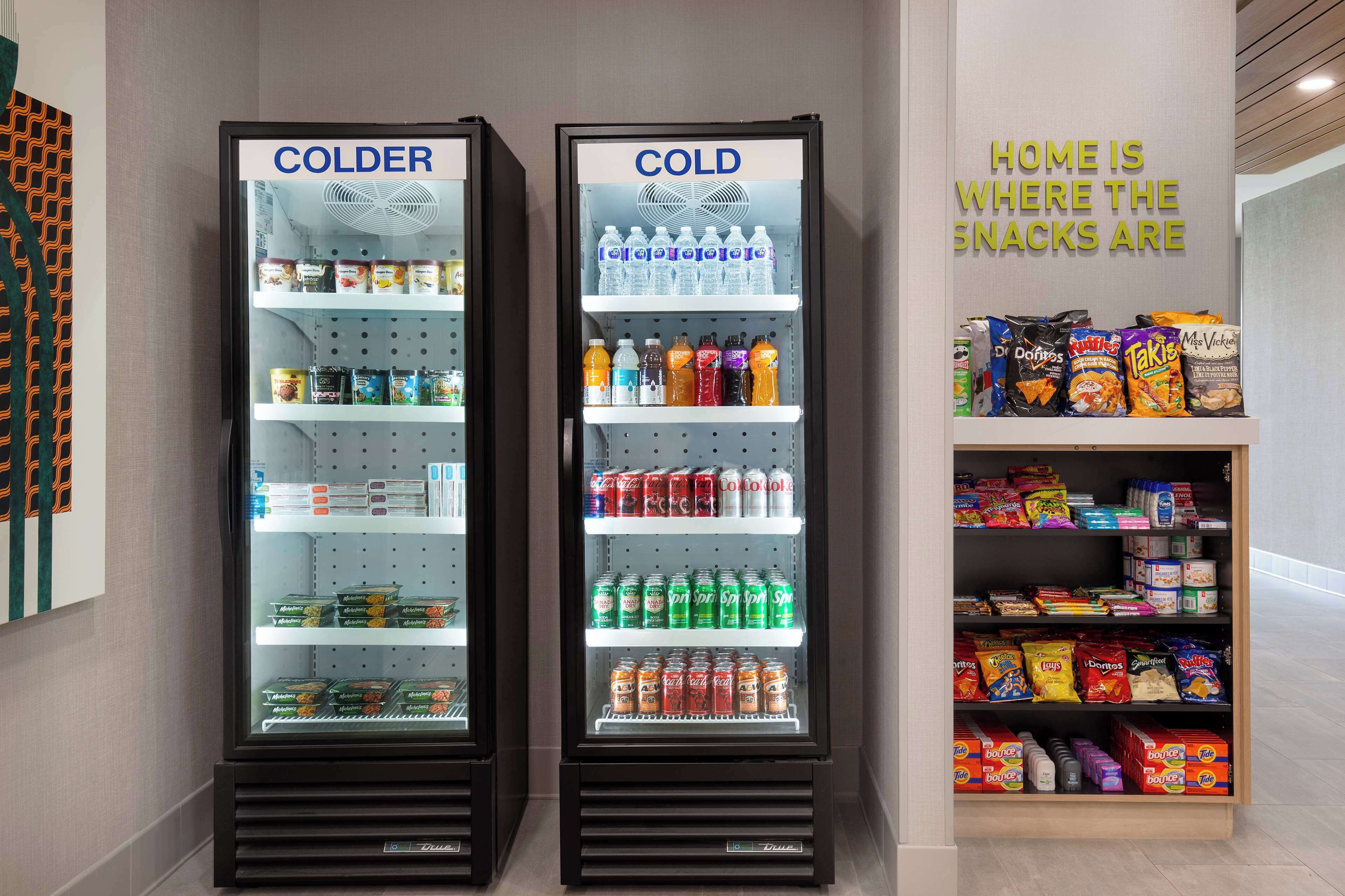 Two refrigerated display cases labeled "COLDER" containing beverages and snacks, with a sign stating "HOME IS WHERE THE SNACKS ARE".
