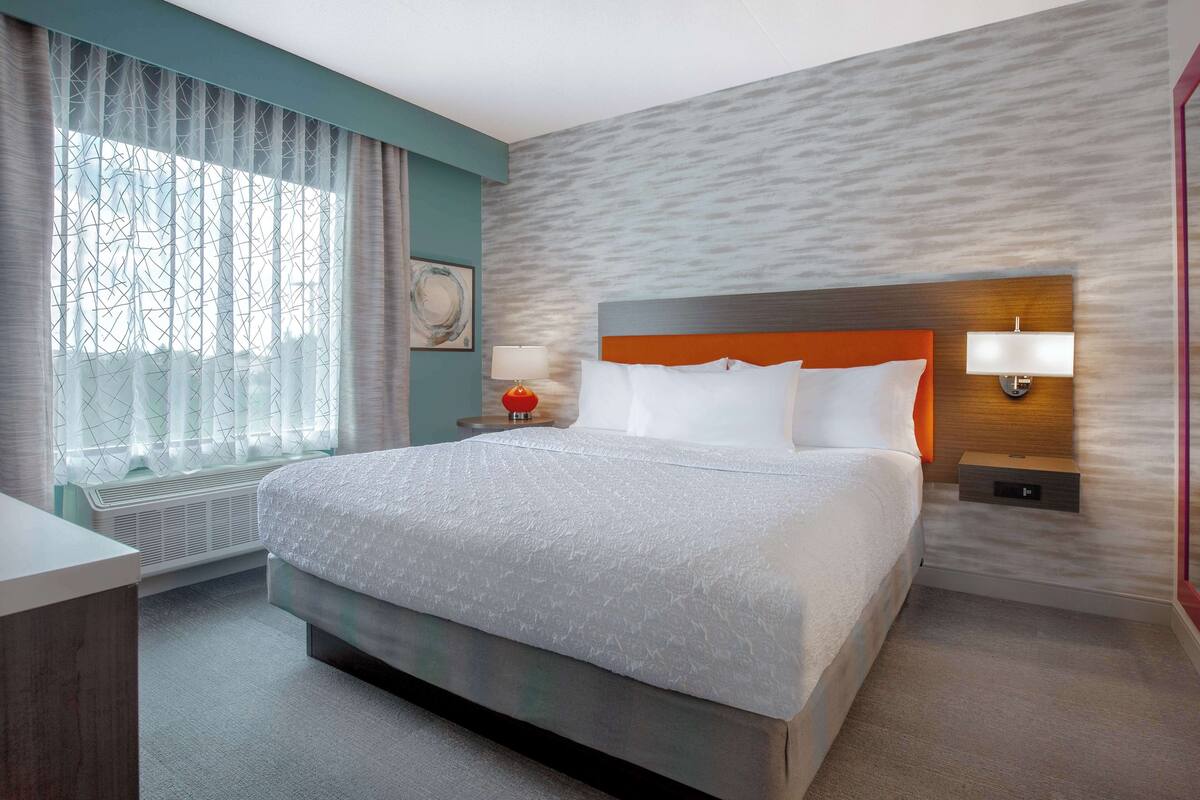 A hotel room with a large bed, a bedside table with a lamp, a wall-mounted light, and a window with sheer curtains.