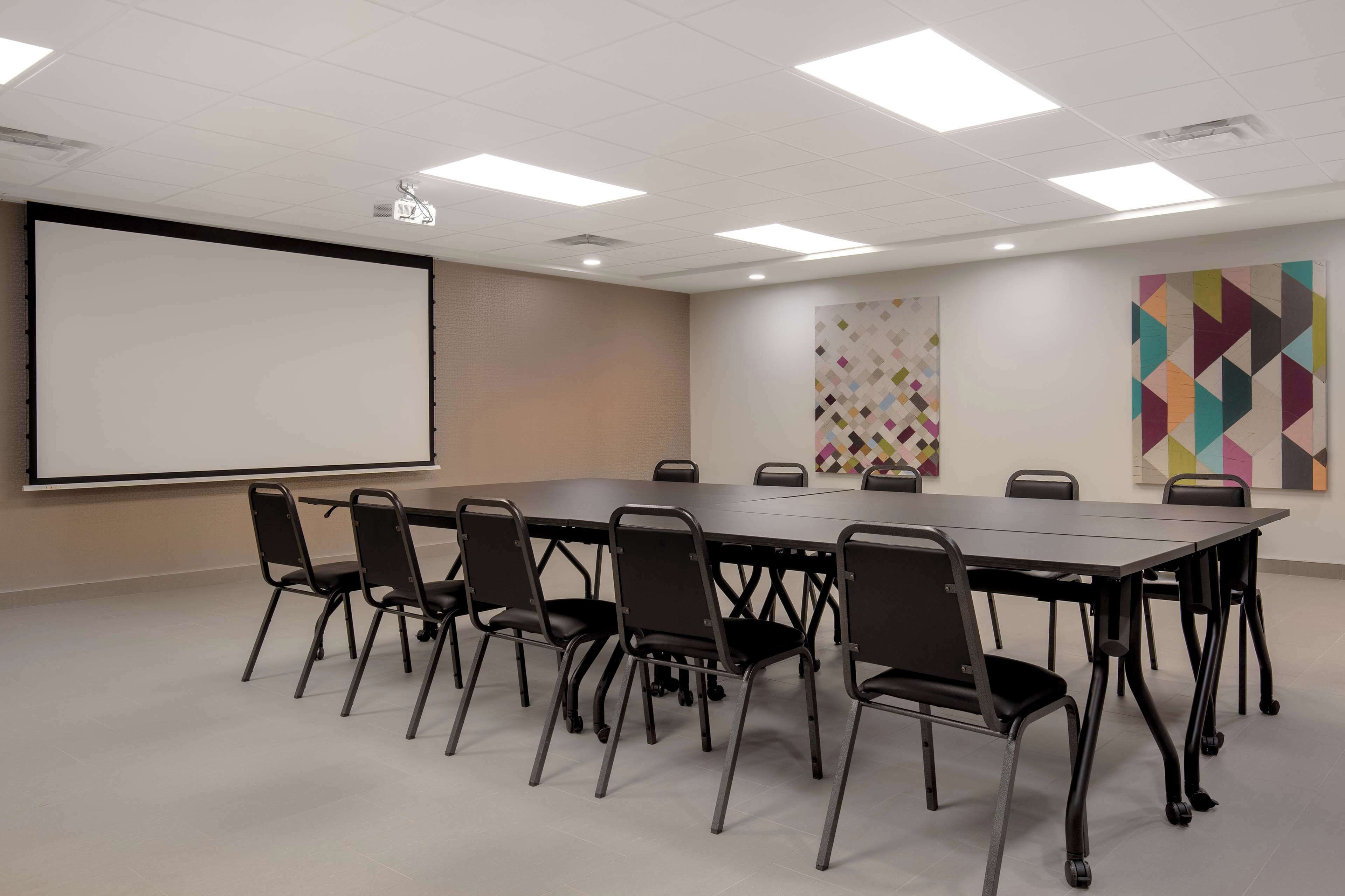 A conference room with a long table, black chairs, a projector screen, and colorful wall art.