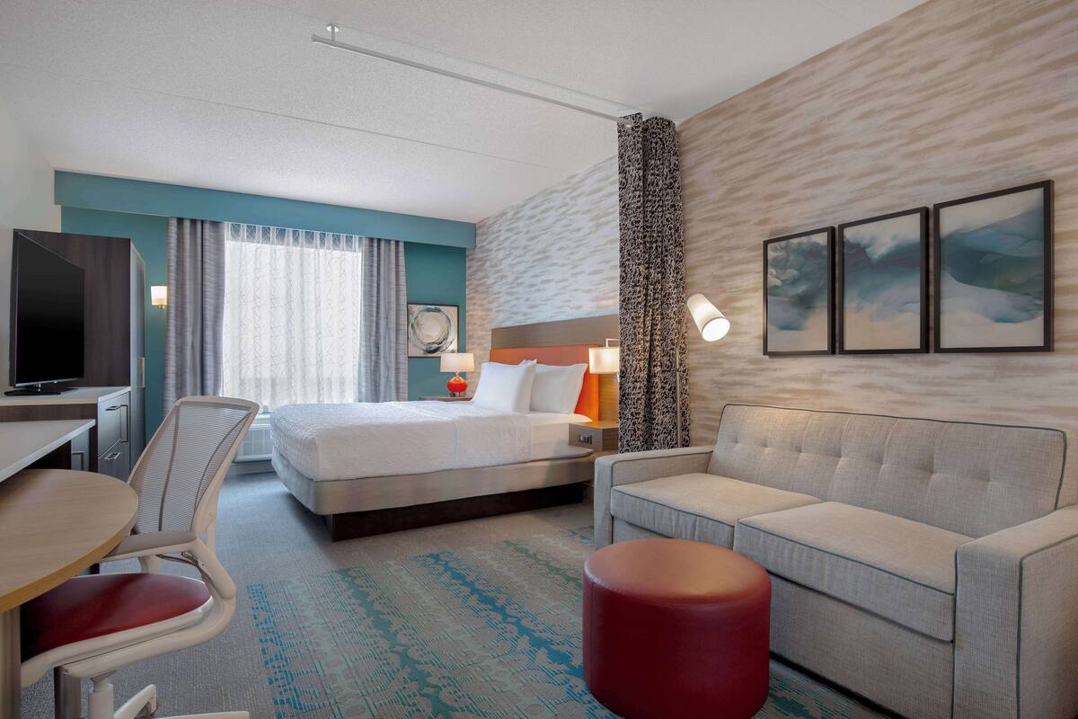 A hotel room with a bed, a sofa, a desk with a chair, a TV, and a window with curtains.
