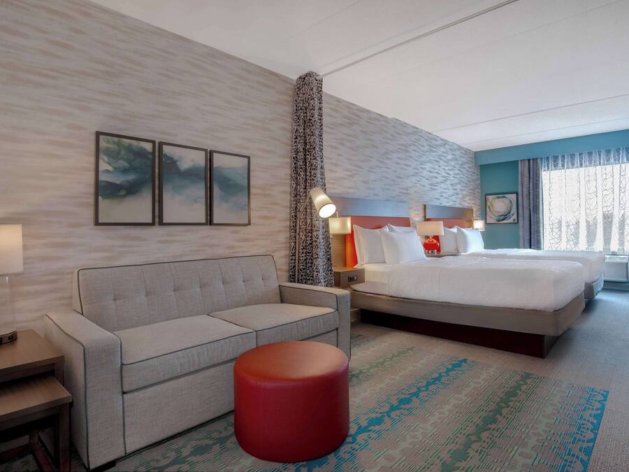 A hotel room with a bed, a sofa, a red ottoman, a nightstand with a lamp, and artwork on the walls.