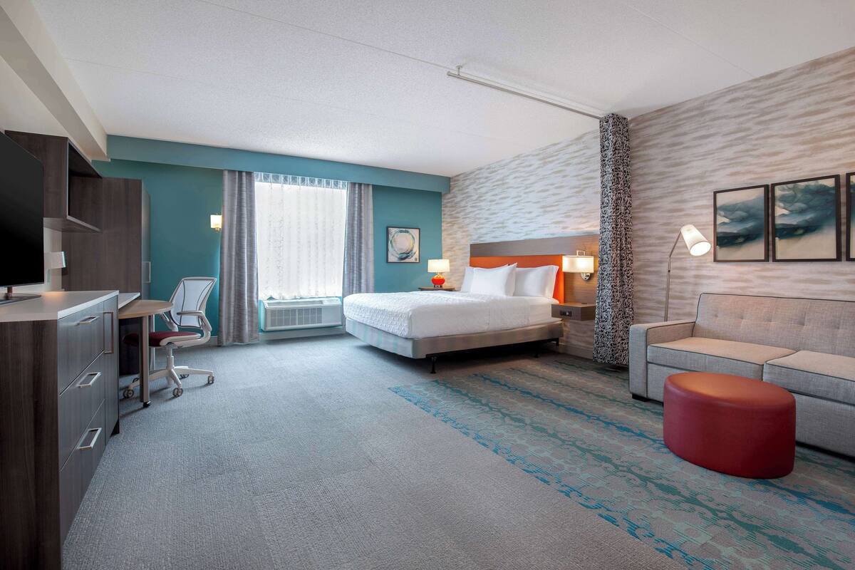 A hotel room with a bed, a desk, a chair, a sofa, and a red ottoman.