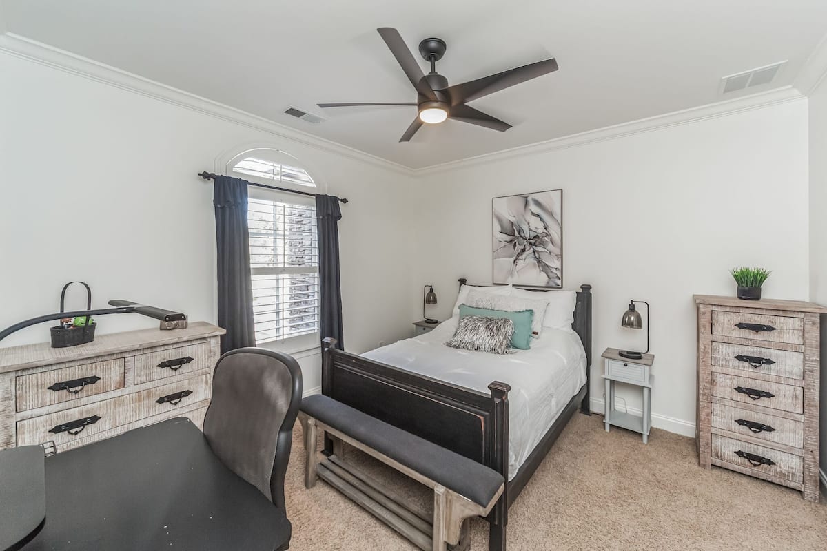A bedroom with a bed, a desk, a chair, a ceiling fan, a window with curtains, a nightstand, a lamp, and a dresser.