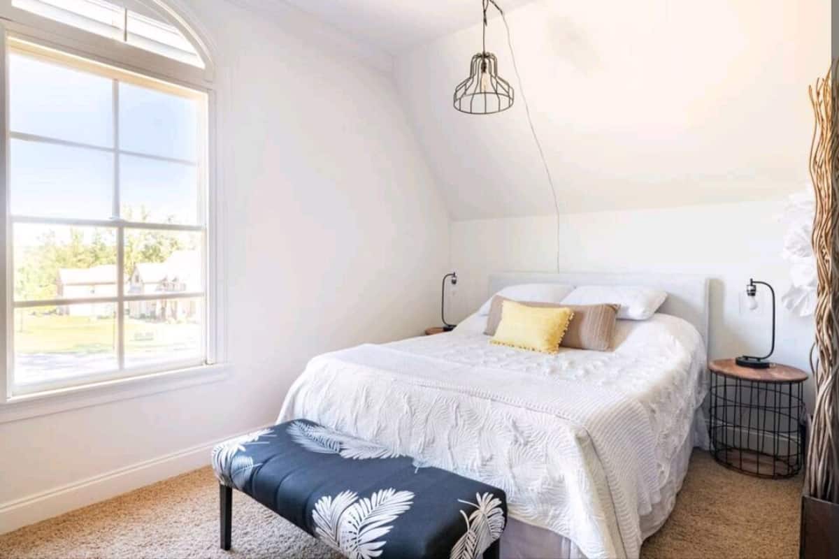 A bedroom with a large window, a bed with white bedding, a bench with leaf patterns, and a hanging light fixture.