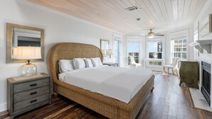 A spacious bedroom with a large wicker bed, a wooden nightstand, a fireplace, and a view of the outside through large windows. - Vast Luxurious Home in Gated Community + Pool (Seacrest)