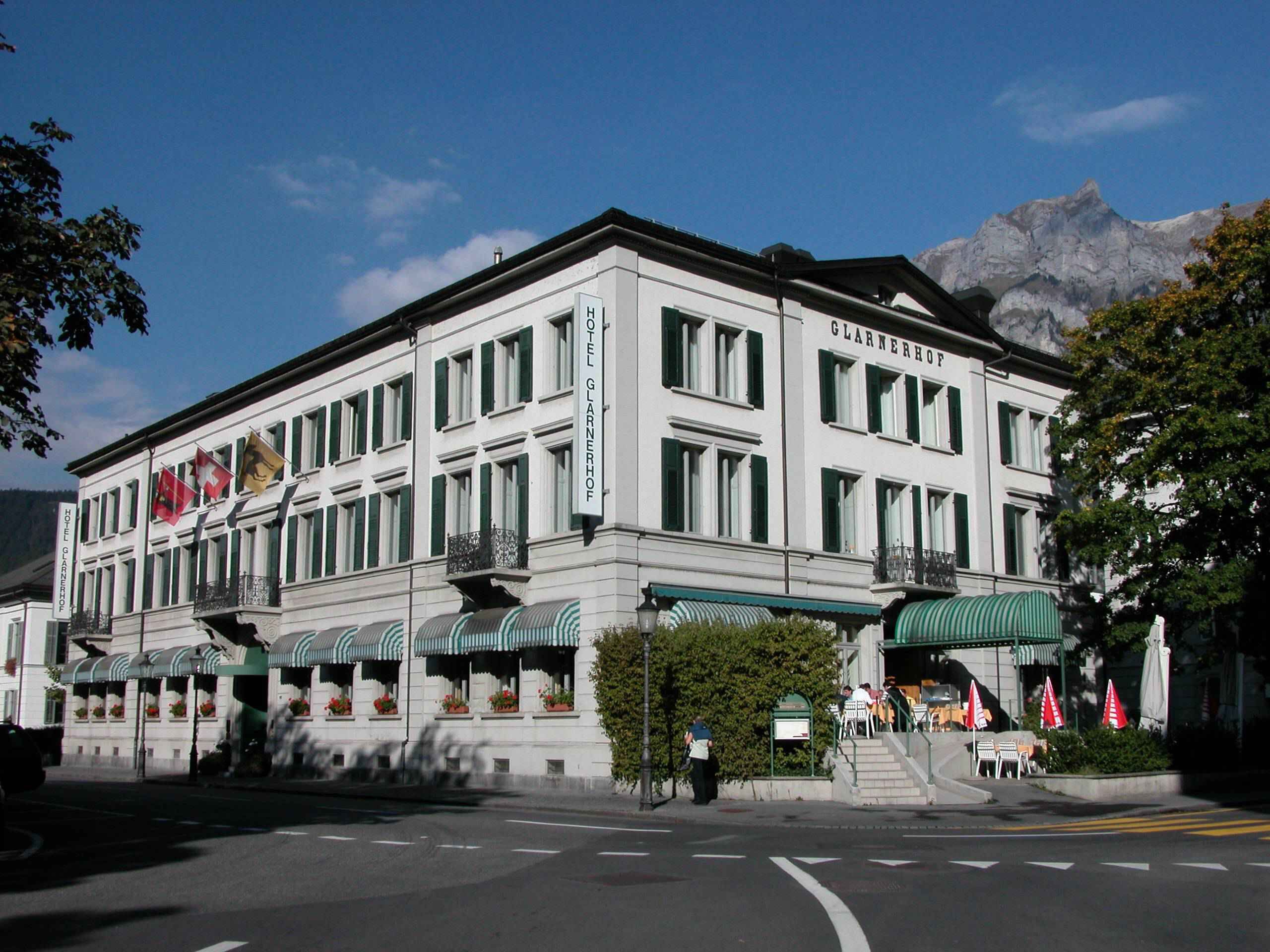 A white building with green shutters, labeled "Hotel Glarnerhof," featuring a balcony and outdoor seating area.