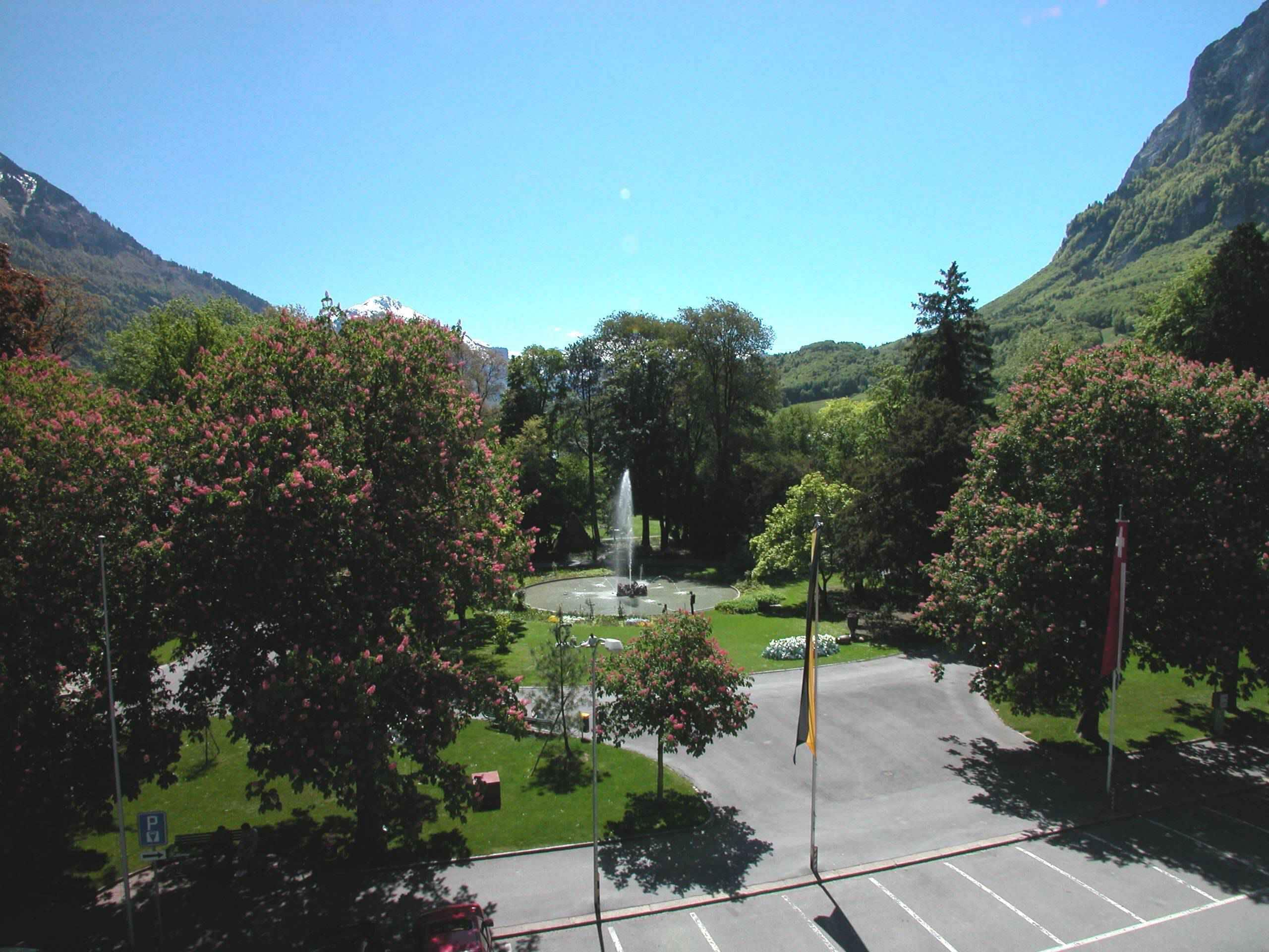 A parking lot with a fountain, surrounded by trees and a mountain backdrop.