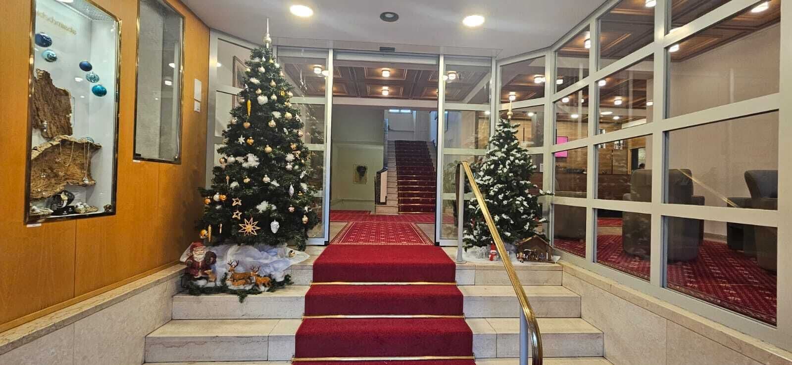 A staircase with red carpet leading to a second floor, flanked by two decorated Christmas trees and a glass wall on one side.