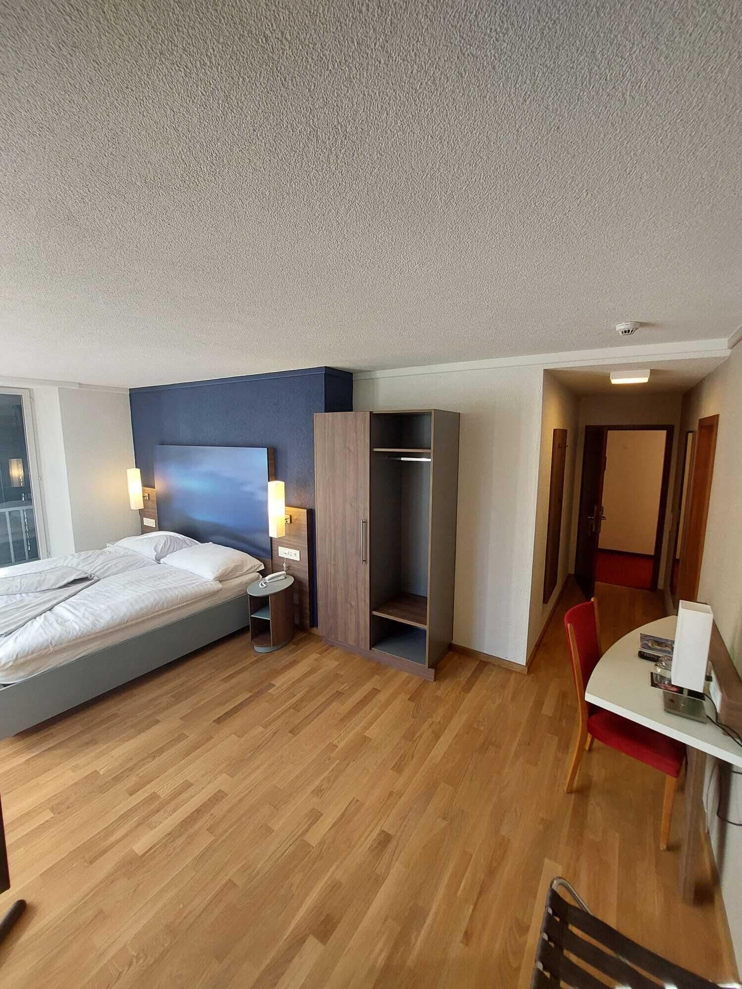 Una habitación de hotel con cama, mesita de noche, lámpara, armario y zona de comedor con mesa y sillas.