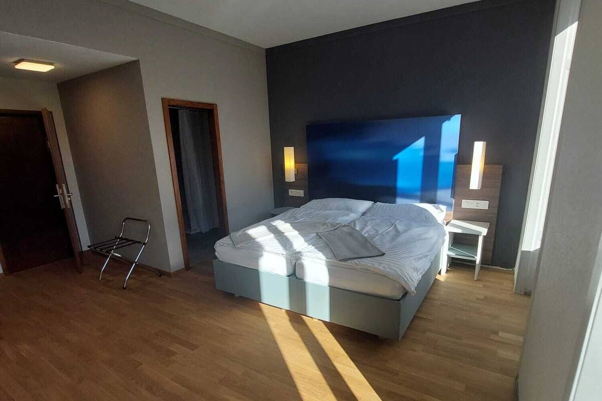 A modern bedroom with a large bed, a wooden floor, a dark accent wall, and a window allowing natural light.