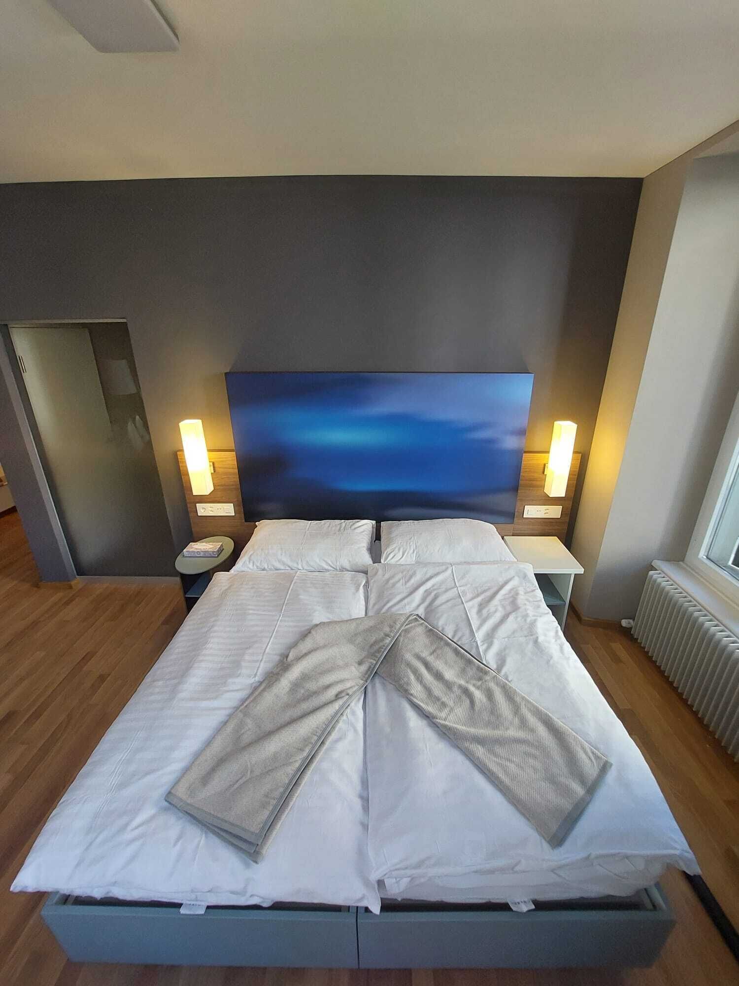 A modern hotel room with a large bed, two bedside tables, a nightstand, and a large window.