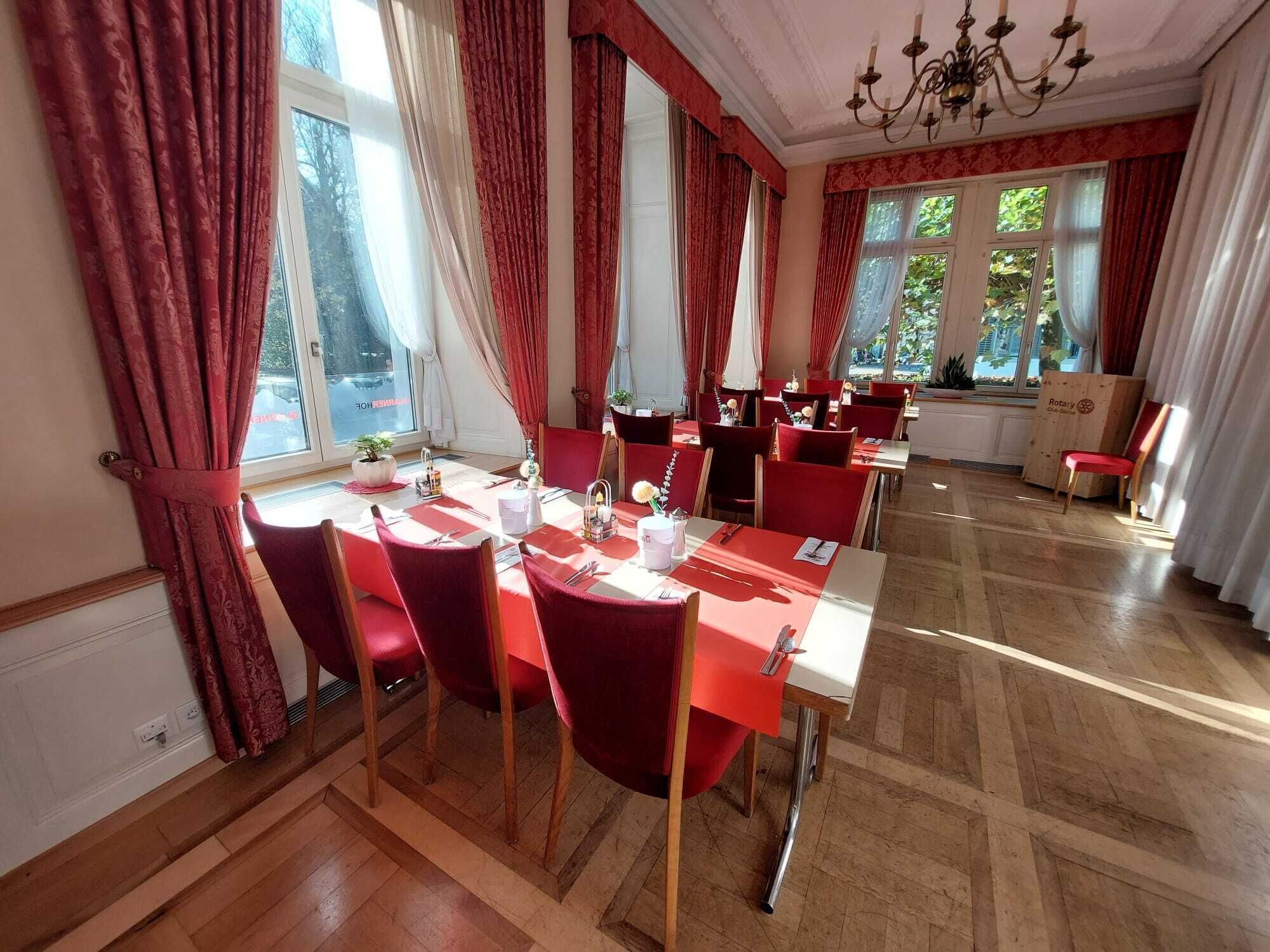 A dining area with red chairs and tables, large windows with curtains, and a chandelier.
