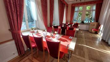 A dining area with red chairs and tables, large windows with curtains, and a chandelier.