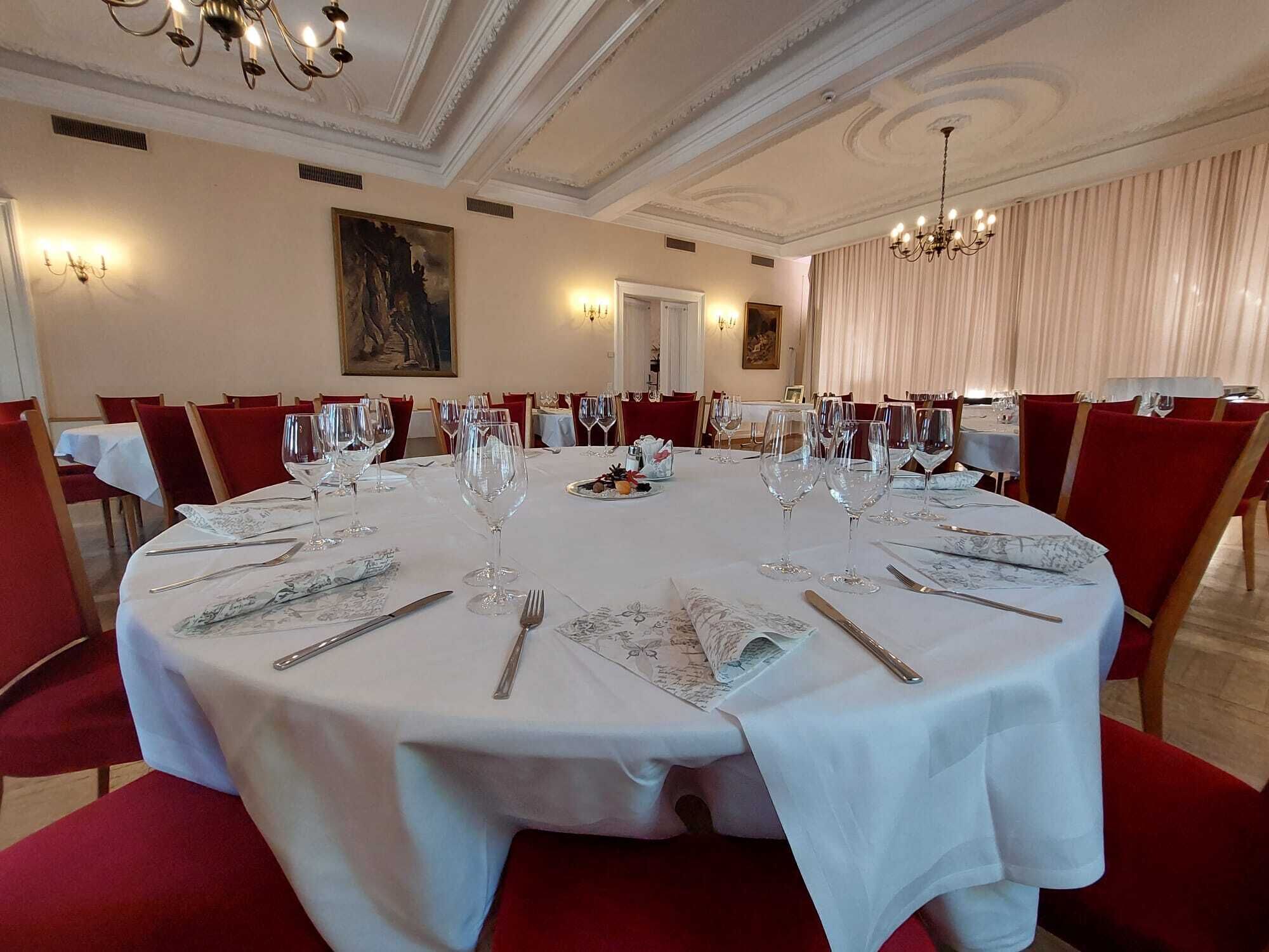 A formal dining room with round tables set for a meal, featuring white tablecloths, red chairs, and elegant chandeliers.