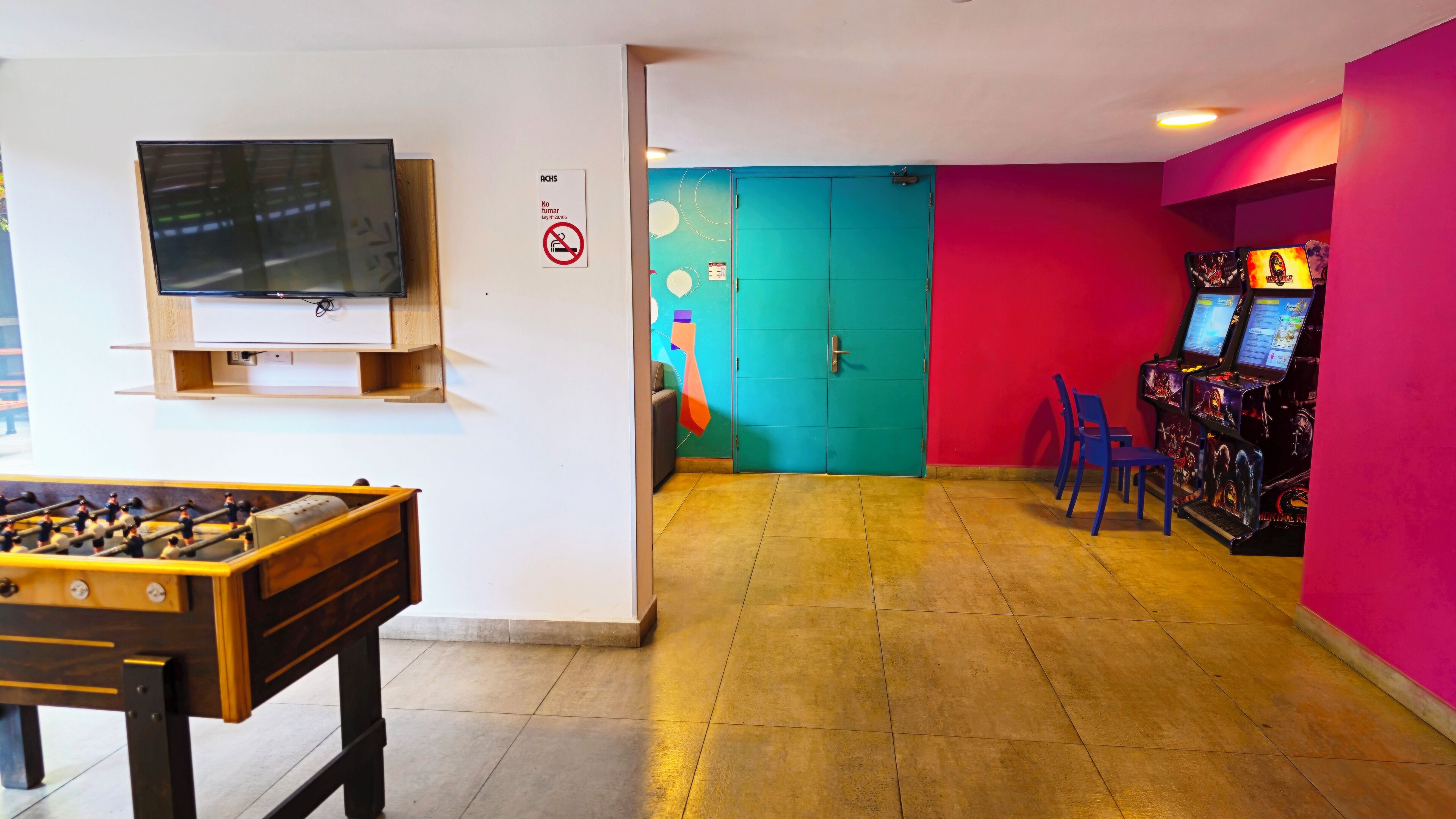 A room with a foosball table, a television mounted on the wall, a teal door, and a red wall with arcade machines.