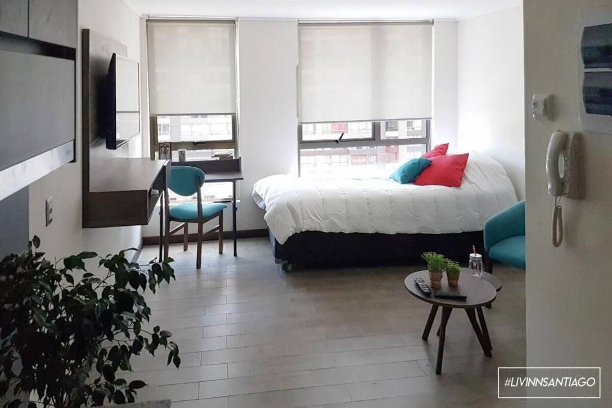 A modern hotel room with a bed, a desk, a chair, and a small table with a plant.
