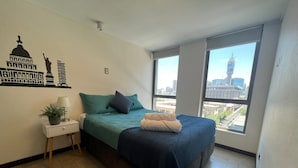 Une chambre à coucher comprenant un lit, une table de chevet, une lampe, une fenêtre donnant sur la ville et un motif décoratif sur le mur.