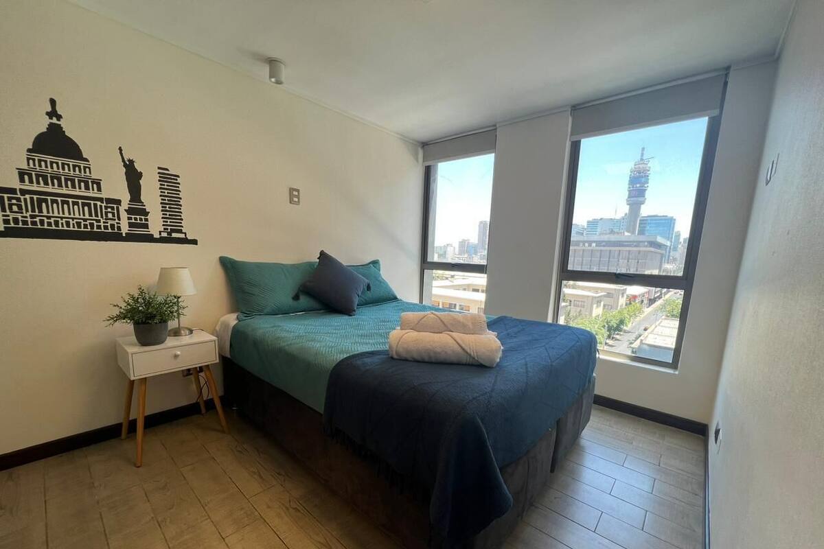 A bedroom with a bed, a nightstand, a lamp, a window with a city view, and a wall decal.