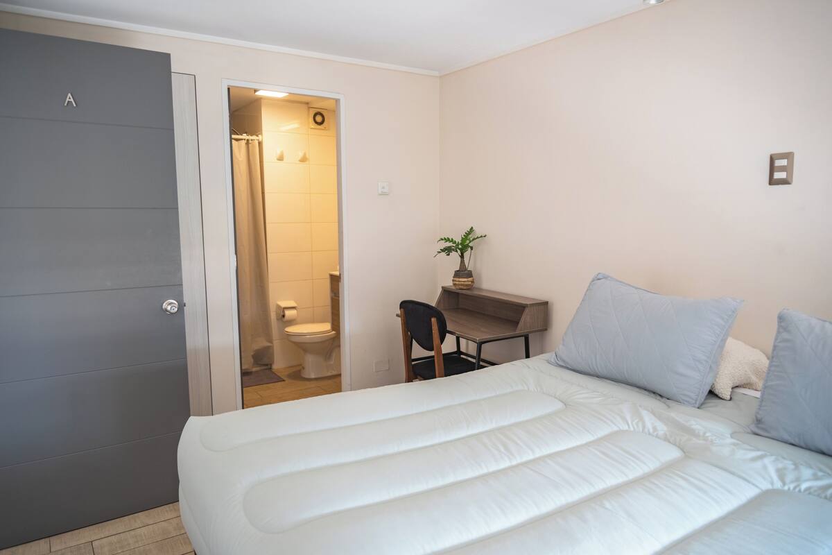 A bedroom with a bed, a desk, a chair, a plant, and a bathroom visible through an open door.