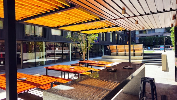 Un espace extérieur aménagé avec des tables et des bancs en bois, un escalier menant à un niveau supérieur, et un bâtiment moderne aux grandes fenêtres en arrière-plan.