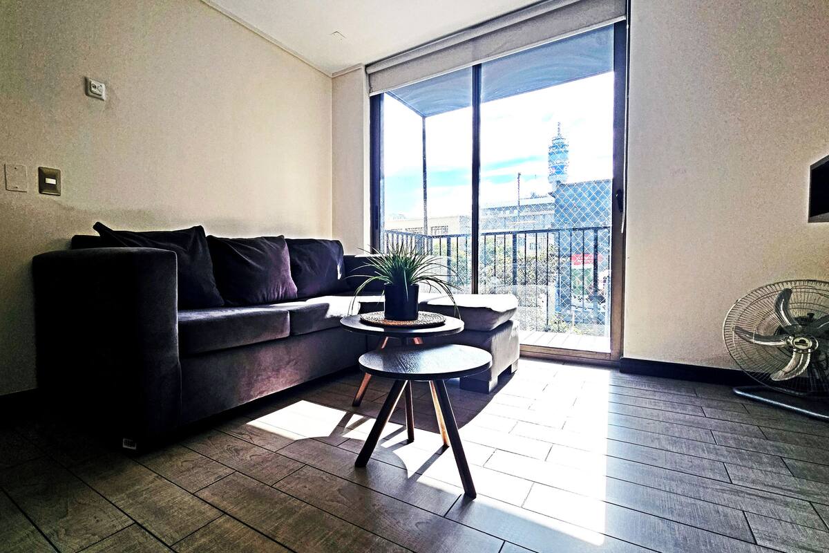 A modern living room with a dark-colored sofa, a small round table with a potted plant, and a large window offering a view of a cityscape.