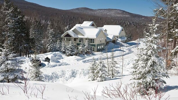 Ein großes, mehrgeschossiges Haus mit schneebedecktem Dach, umgeben von schneelastigen Bäumen und einer Berglandschaft im Hintergrund.