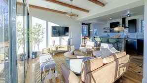 A spacious living area with a large sofa, a coffee table, and a dining area with chairs. There is a kitchen visible in the background with modern appliances and a large window.