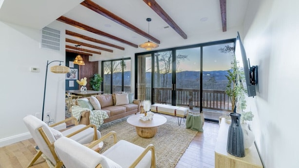 A spacious living room with a large sliding glass door leading to a balcony, wooden beams on the ceiling, and a round coffee table.