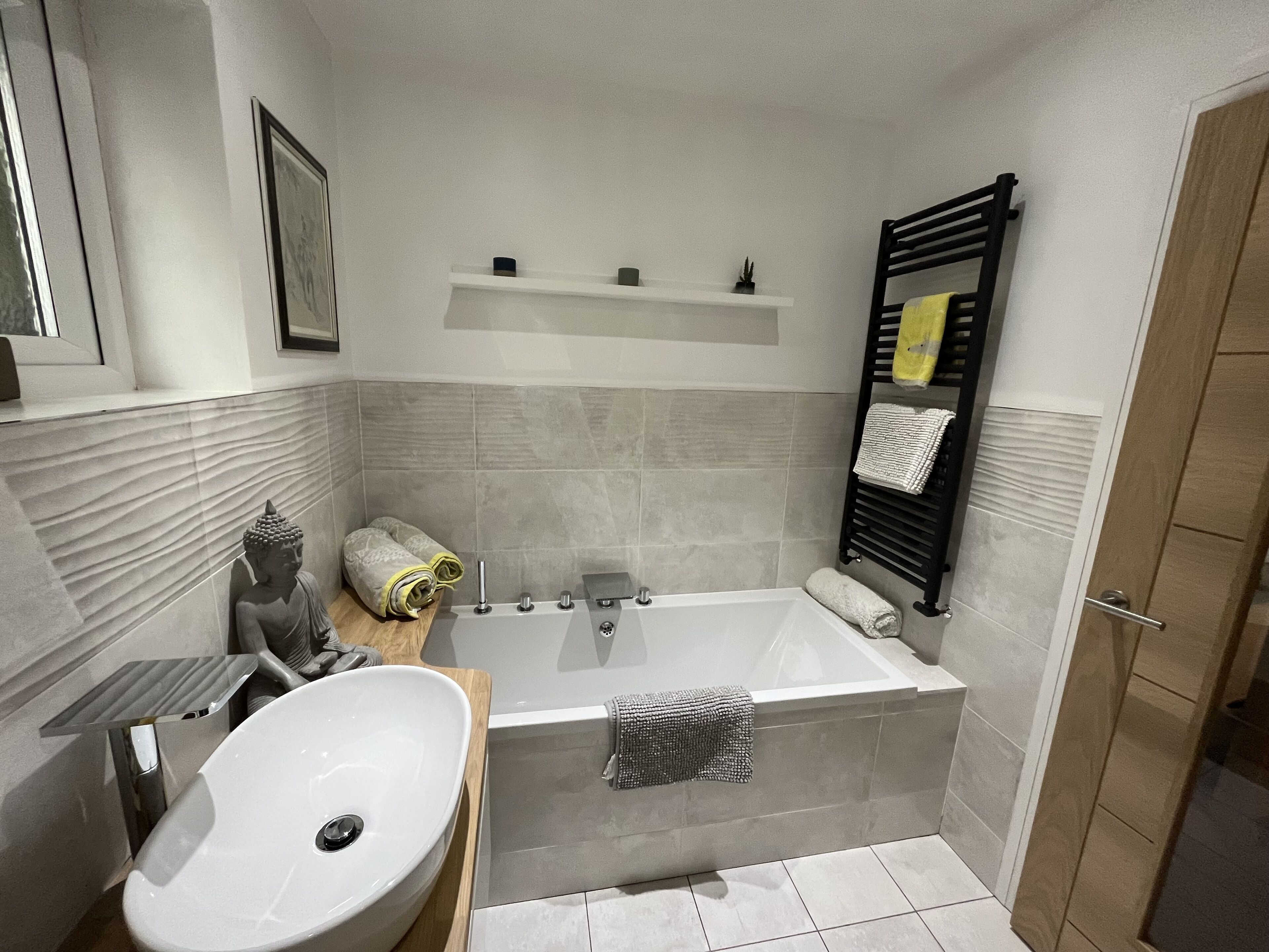 A modern bathroom with a bathtub, a sink, and a toilet. There is a shelf above the bathtub and a towel rack on the wall.