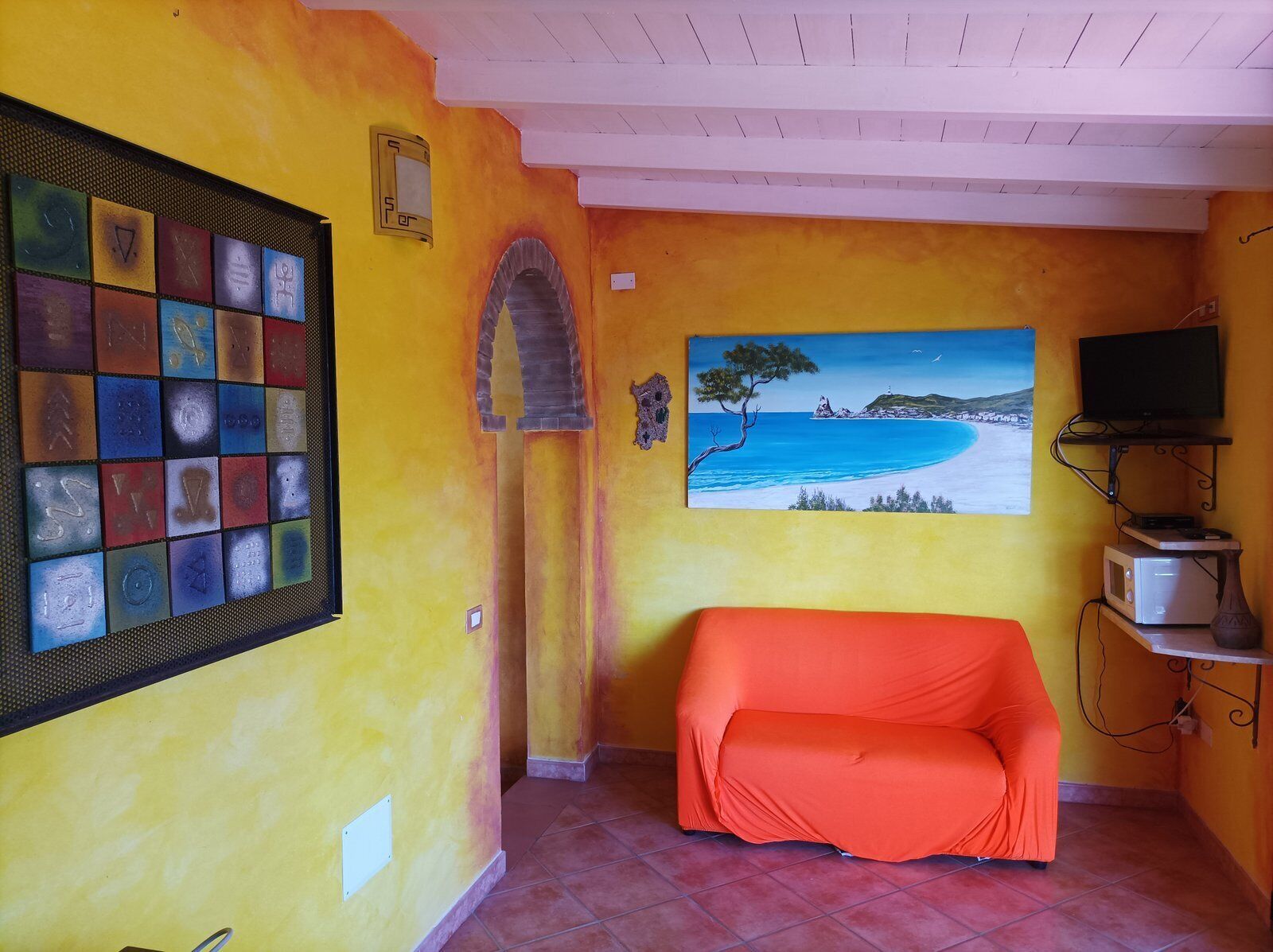 Una habitación con una pared amarilla, un cuadro de playa, un televisor en un estante y un sofá.