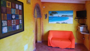 Ein Zimmer mit einer gelben Wand, einem Strandbild, einem Fernseher auf einem Regal und einem Sofa.